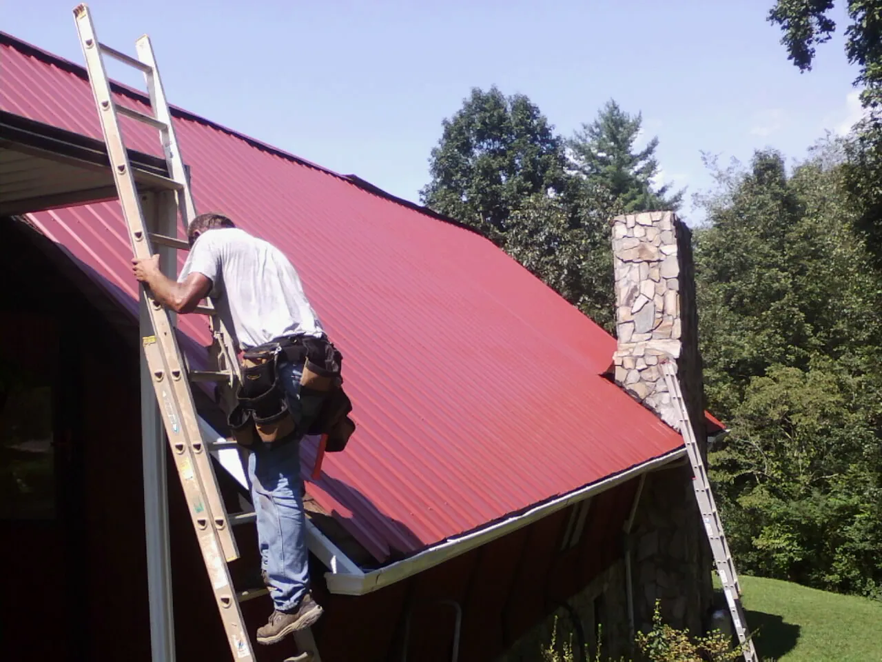 Roofer on a ladder beside a red metal roof and stone chimney — Soffit & Fascia Repair by Roofing Brothers in Yorklyn